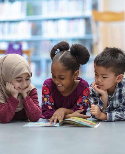 Three young children (left to right: a young girl in a hijab, a girl of colour with her hair in buns and an Asian boy with short hair) are sitting at a table, smiling. They are looking at an open book on the table.