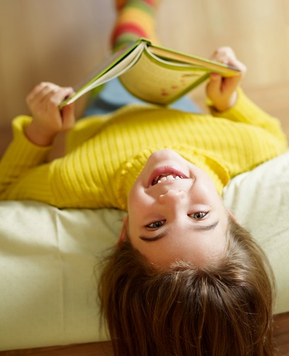 A young girl is hanging upside down on a bed, holding a book. She is smiling at the camera.
