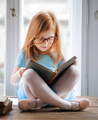 A young girl with red hair sits cross-legged on a window sill. She is holding a book and wearing glasses, and looking down at the book.