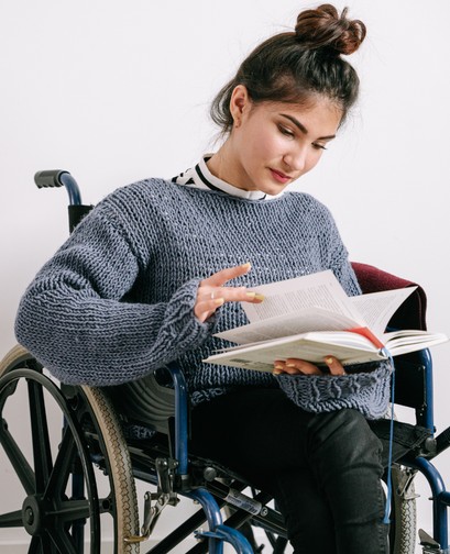 A young woman in a wheelchair is reading a book, looking thoughtfully at it.
