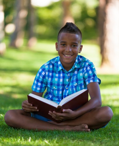 A young boy of colour is wearing a plaid shirt, sitting cross-legged on the grass and holding a book. He is smiling at the camera.
