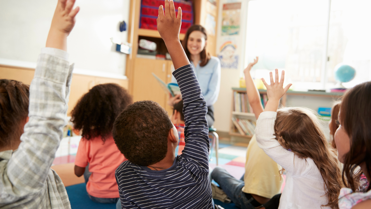 A teacher sits smiling in the background among a group of children sitting with their hands up in the foreground.