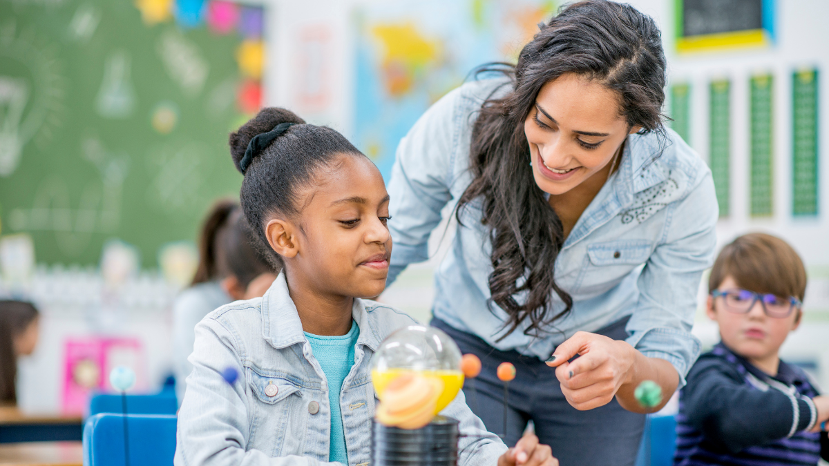 A teacher leans over a student and smiles as she sees the molecule model she has been working on.