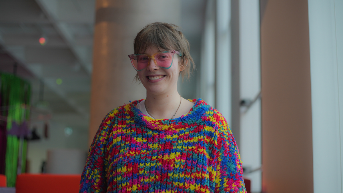 A photo of the author, Alex Creece. She wears a colourful woollen jumper and glasses.