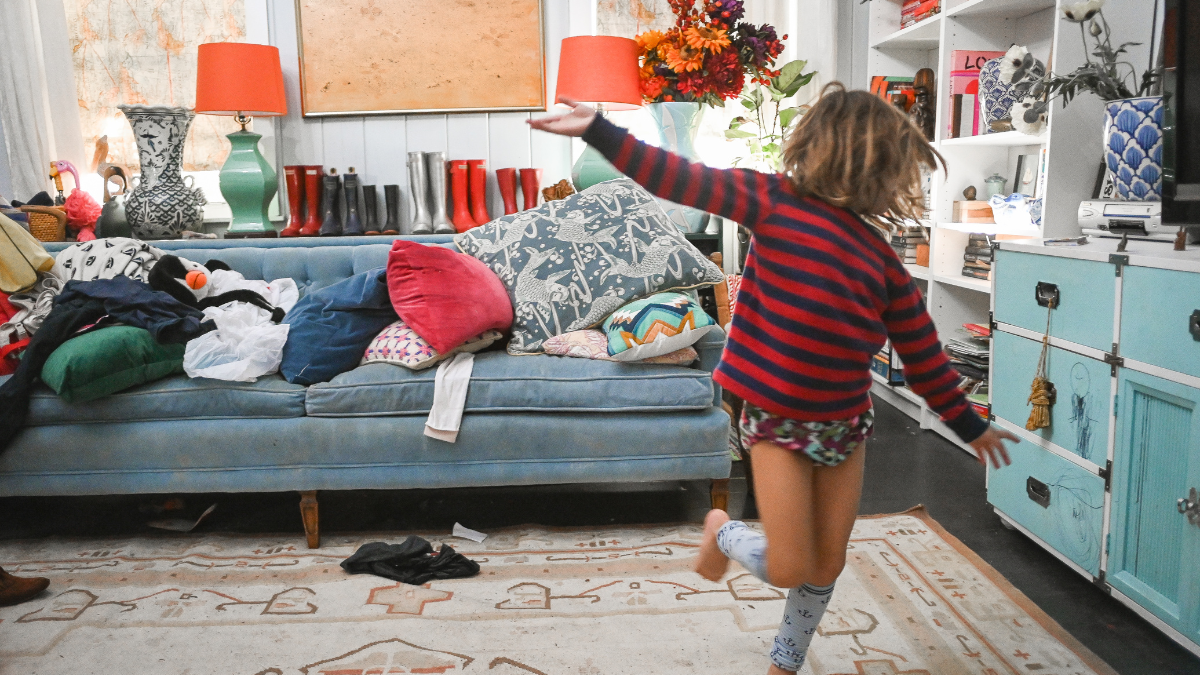 A kid runs around a messy living room with their arms oustretched.