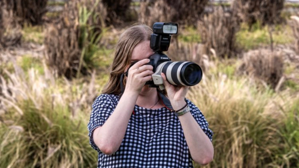 An image of Jessica, a young woman with long, dark blonde hair wearing a black and white gingham dress whilst standing in a grassy field. She is holding a professional DSLR camera and is looking through the lens.
