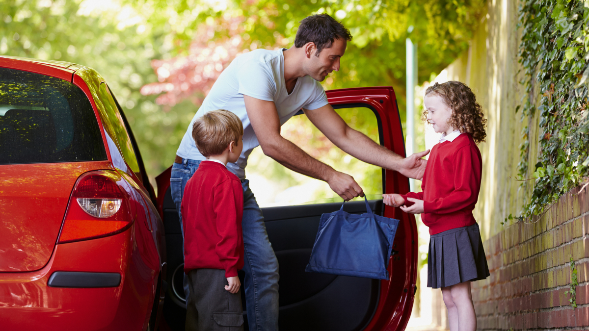 A father offers his daughter her school bag as she and her brother, dressed in school uniforms, get out of the car.