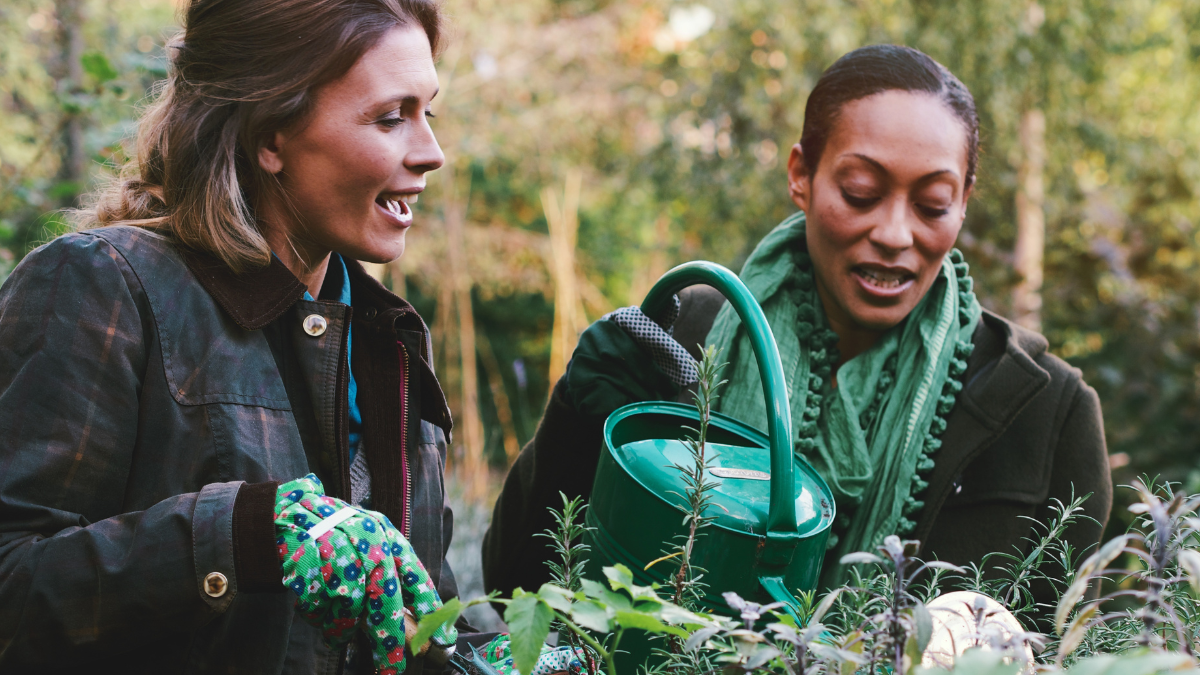 A photo of two adult female friends helping each other work in a community garden