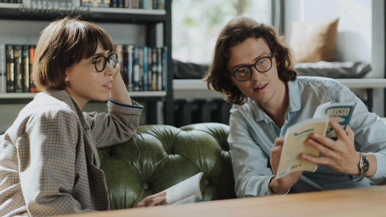 Two young adults sit at a table, discussing a book.