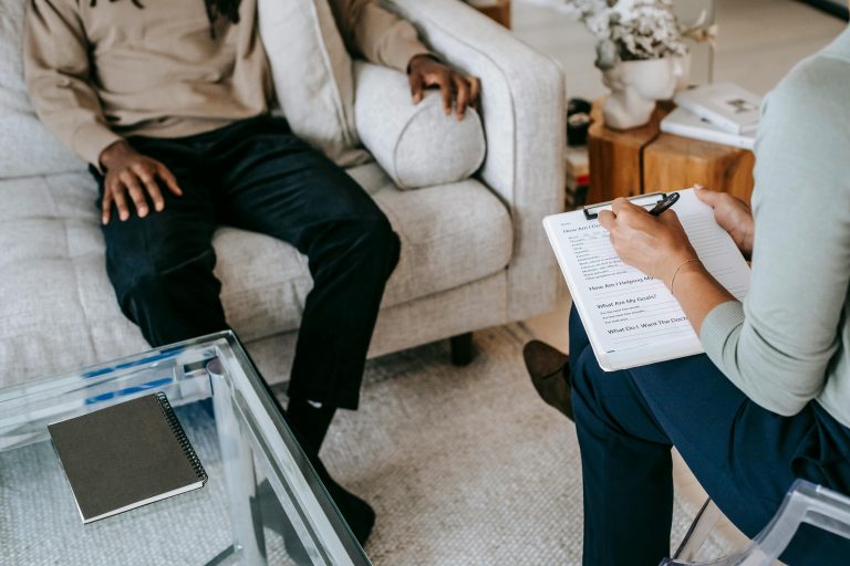 Photo of a man sitting on a sofa across from a psychologist who is filling in a form.