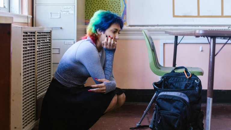 A teenager with multi-coloured hair sits crouched, covering her mouth in a classroom.