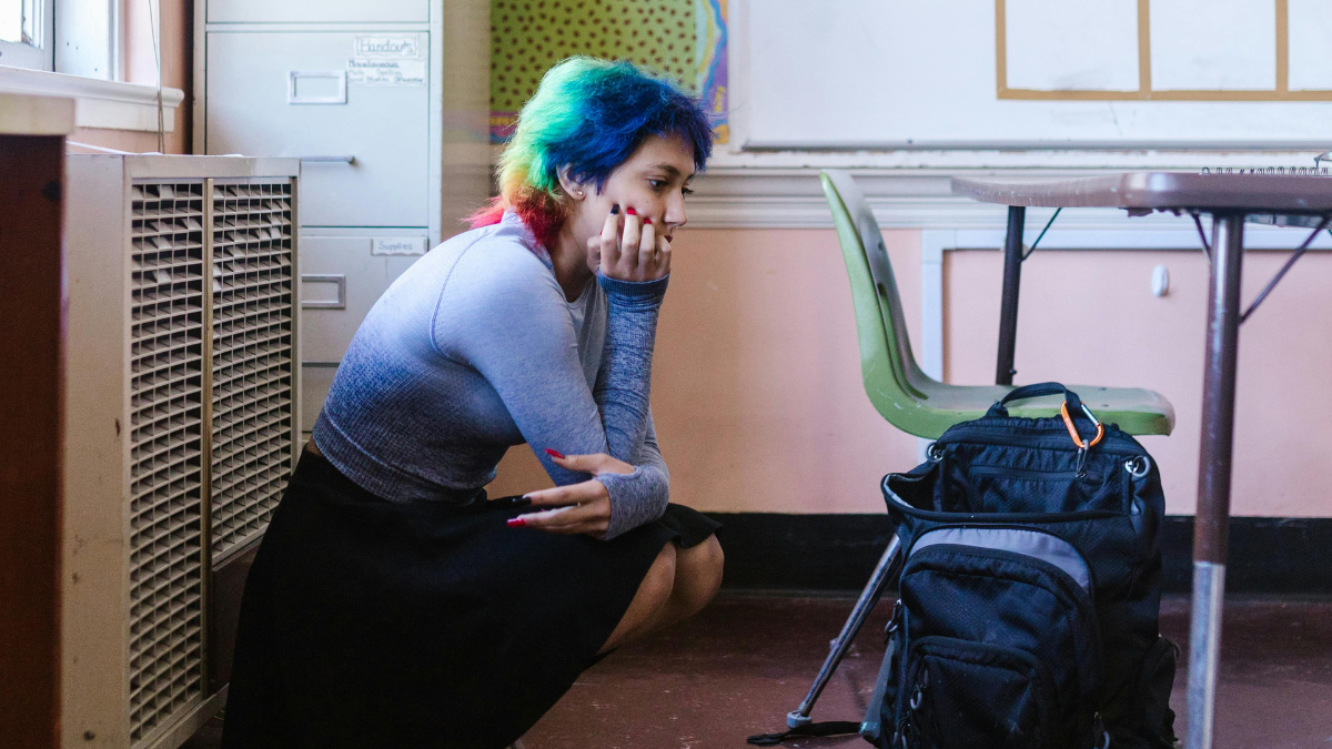 A teenager with multi-coloured hair sits crouched, covering her mouth in a classroom.