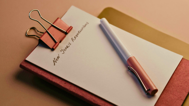 A pen lies on a clipboard with a piece of paper with the title 'New Year's Resolutions'