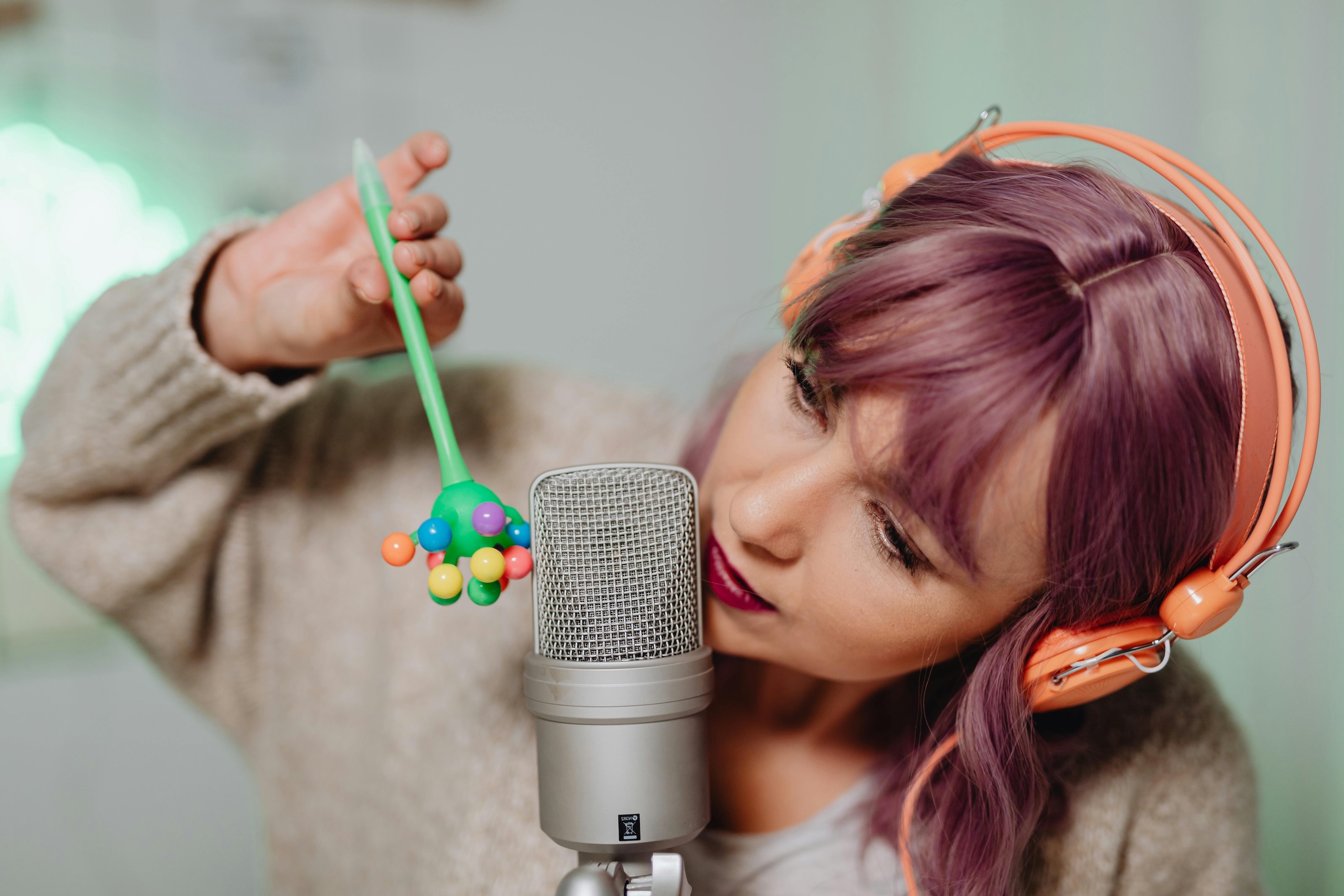 A photo of a woman wearing orange headphones, by a microphone. She is playing with a stim toy.